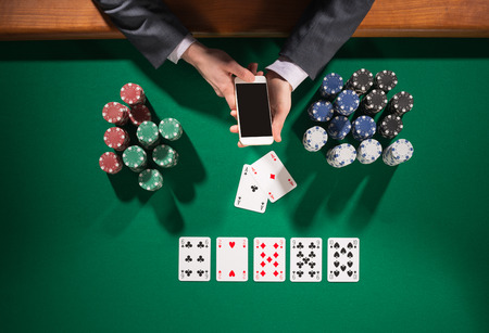 poker: Elegant male poker player using a smartphone cards and chips stacks all around green table top view