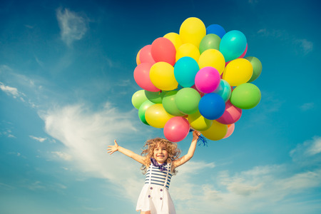 balloon: Happy child jumping with colorful toy balloons outdoors. Smiling kid having fun in green spring field against blue sky background. Freedom concept
