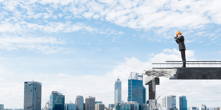 Young engineer in suit and helmet looking in binoculars while standing on broken bridge with cityscape on background 3d rendering Stock Photo