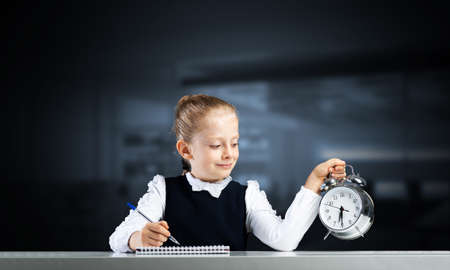 目覚まし時計を保持している小さな女の子の笑顔。勉強して学校のコンセプトに戻る時間です。手にノートとペンで机に座っている幸せな女子高生。時間管理。学校での研究と教育。の写真素材・画像素材 Image 159384401