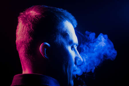 A view from the back of a boy’s head around in a shirt smoking a cigarette and exhaling multi-colored smoke of blue to the right side from himself on a black isolated background. harm to health.