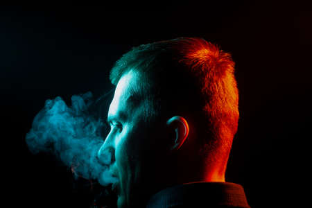 A view from the back of a man’s head around in a shirt smoking a cigarette and exhaling multi-colored smoke of green to the left side from himself on a black isolated background. harm to health of man