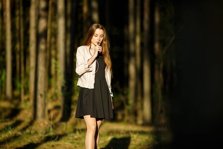Vape teenager. young cute girl in casual clothes smokes an electronic cigarette outdoors in the forest at sunset in summer. bad habit that is harmful to health. vaping activity.