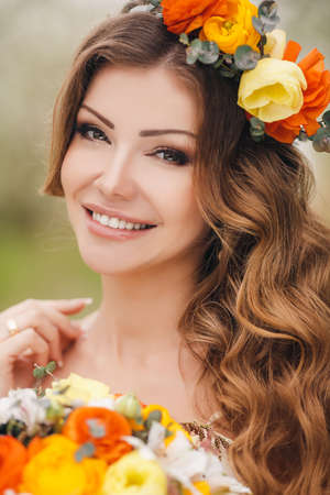 明るい花の花輪を身に着けている手で彼の頭の上のウェディング ドレスで幸せな花嫁がブライダル ブーケ 美しい髪 長い巻き毛の白い花を持つ緑豊かな春の庭でポーズを保持しています の写真素材 画像素材 Image