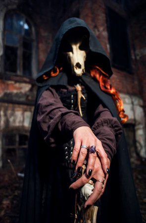 Scary red-haired witch with a staff in a black cloak with a hood and a skull at night against the background of an old sinister castle. focus on the foreground