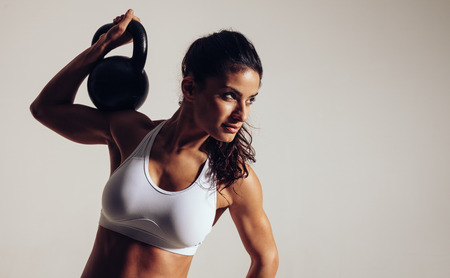 Focused young woman doing crossfit workout with kettle bell on grey background. Woman in sportswear looking away at copy space while crossfit workout session. Фото со стока