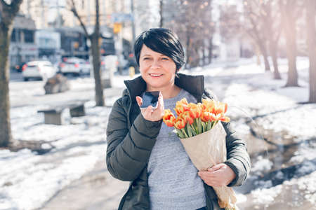 Middle Age Brunette Woman Walking On Spring Street With Yellow Tulips Flowers And Talking On Audio Chat Phone. New Social Media App. Speaker In Live Stream On Smartphone. Using Technology.