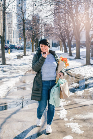 Middle Age Brunette Woman Walking On Spring Street With Yellow Tulips Flowers, Shopping Bags And Talking On Audio Chat Phone. New Social Media App. Speaker In Live Stream On Smartphone.