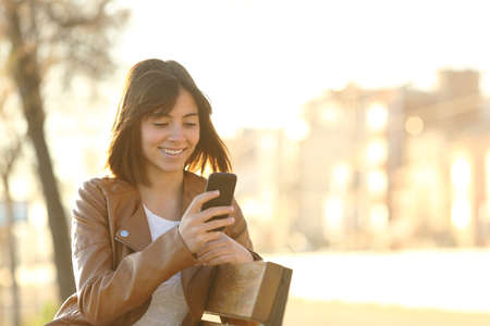 Happy girl using a smart phone in a city park sitting on a bench