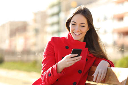 Girl texting on the smart phone sitting in a park wearing a red jacket and sitting in a bench in a park Stock Photo