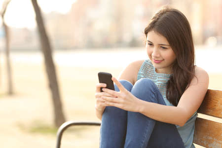 Teen girl using a smart phone and texting sitting in a bench of an urban park Stock Photo