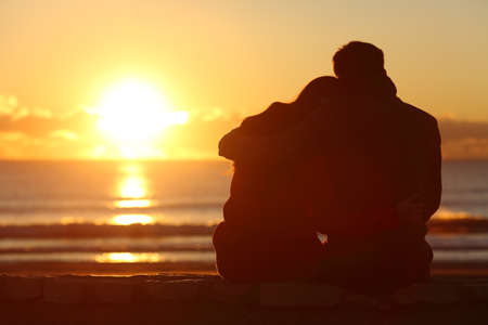 Vista posterior de una silueta de pareja mirando el sol al atardecer en la playa en invierno con una luz cálida - 64631613