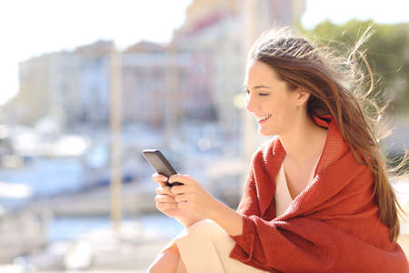 Girl sitting using a smart phone texting messages on line in a port of urbanization with the sea in the background and the wind moving her hair Stock Photo