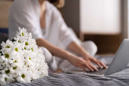 Young Woman Sitting On The Bed Wearing Pajamas, With Pleasure Enjoying White Flowers, Chatting Using Laptop