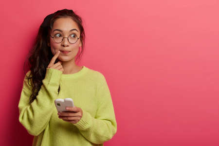 Photo of pensive surprised woman keeps index finger near lips, looks aside, thinks about message content, holds mobile phone, wears transparent glasses and green sweater, isolated on pink wall