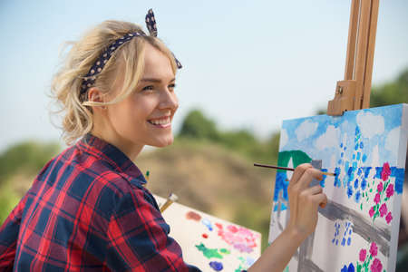 Beautiful blonde woman artist in a playful mood dressed in a plaid shirt. She paints a picture with a brush and paints nature Фото со стока