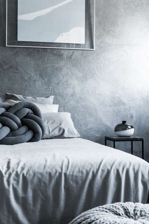 Decorative black and white vase on table next to bed with knot pillow in simple bedroom with poster