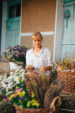 Beautiful Adult Woman In A Shirt Sits Among Baskets Of Dried Flowers And Makes Beautiful Holiday Bouquets. Small Business To Create Bouquets In The Country.