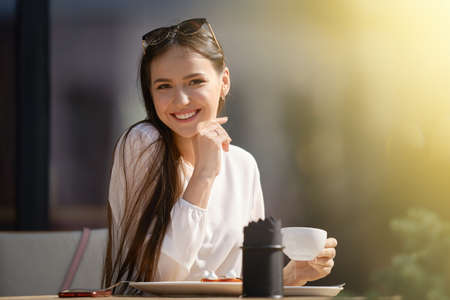 Cute young woman with cup of cappuccino in hand looks straight and smiles