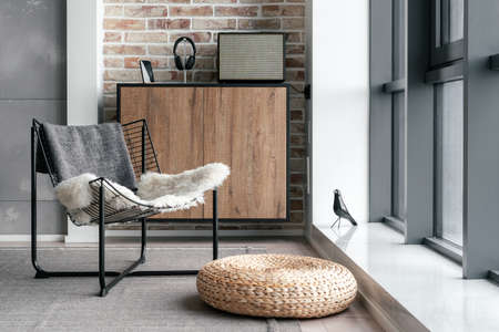 Espace salon confortable près d'une fenêtre panoramique avec chaise en métal, pouf en paille et équipement moderne pour écouter de la musique sur une commode contre un mur de briques. design d'intérieur de style loft à l'hôtel ou à la maison