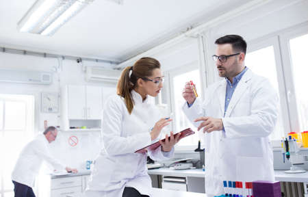 Two young lab technicians scientists talking experimenting in laboratory. man and woman doctors holding vial clipboard doing research.