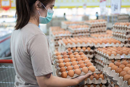 Femme asiatique portant un masque facial, choisissant des œufs de poule frais lors de l'achat de nourriture, les gens paniquent en achetant et en accumulant pendant le covid-19, épidémie de coronavirus. fille se préparant à une pandémie de virus pathogène Banque d'images