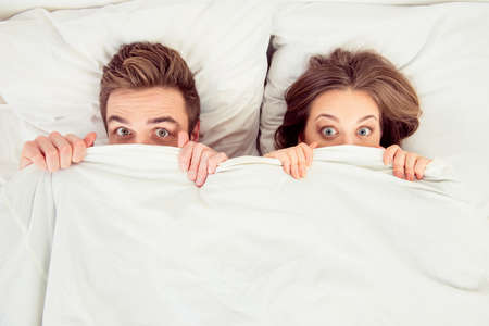 Surprised funny couple in love lying in the bed hiding themselves under blanket - 55107619