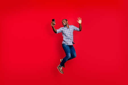 Full length body size of nice funny crazy handsome cheerful optimistic guy wearing checkered shirt holding in hands cell waving hi hello isolated on bright vivid shine red background