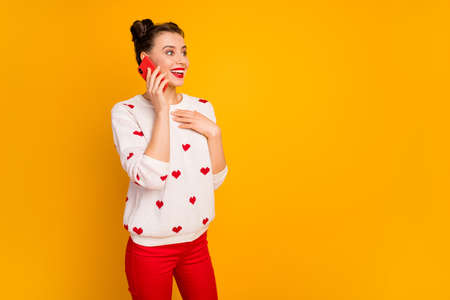Photo of funny energetic lady hold telephone talk best friend tell romantic date story inspired wear, hearts pattern white pullover red trousers isolated yellow color background Stock Photo