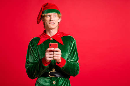 Photo of scared elf guy hold smartphone look copyspace bite lips wear green headwear isolated on bright red color background