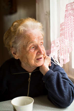 sad lonely: An elderly lady sitting near the window in the kitchen.