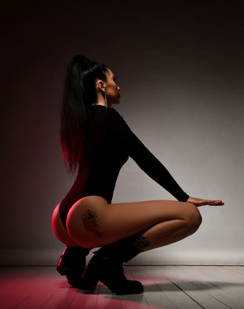 Young slim brunette woman in black thongs bodysuit sits squatting back to camera showing tattoo on