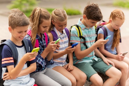 primary education, friendship, childhood, technology and people concept - group of happy elementary school students with smartphones and backpacks sitting on bench outdoors Stock Photo - 65551235 primary education, friendship, childhood, technology and people concept - group of happy elementary school students with smartphones and backpacks sitting on bench outdoors Stock Photo - 65551235