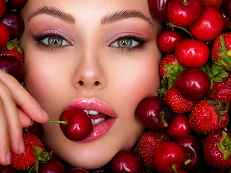 Young girl with bright makeup and a berry background. beautiful caucasian woman biting a berry. attractive woman tasting cherry. beautiful woman with fashion makeup and a vivid background. Banco de Imagens