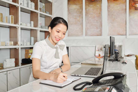 Pretty smiling Vietnamese spa salon receptionist taking notes in planner - 128554217