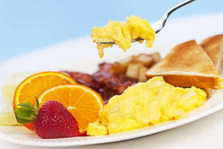 Scrambled eggs on a fork above breakfast plate with fruits toast and bacon - 13306541