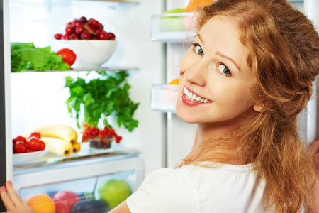 Heureuse femme debout au réfrigérateur ouvert avec des fruits, des légumes et des aliments sains