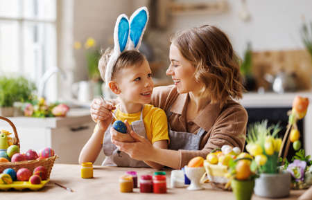 Une jeune mère aimante enseigne à son petit fils heureux à décorer des œufs de pâques tout en étant assis dans la cuisine