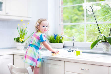Cute curly toddler girl in a colorful dress washing dishes, cleaning with a sponge and playing with foam in the sink in a beautiful sunny white kitchen with a garden view window in a modern home