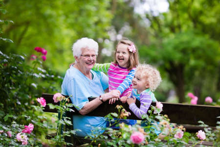 Happy senior lady playing with little boy and girl in blooming rose garden. Grandmother with grand children sitting on a bench in summer park with beautiful flowers. Kids gardening with grandparent. Фото со стока