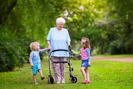 Happy senior lady with a walker holding hands of little boy and girl. Grandmother with grand children enjoy a walk in summer park. Kids supporting disabled grandparent.