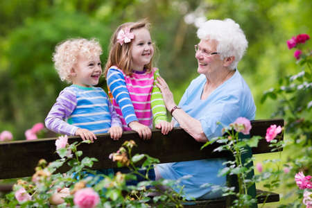 Happy senior lady playing with little boy and girl in blooming rose garden. Grandmother with grand children sitting on a bench in summer park with beautiful flowers. Kids gardening with grandparent. Фото со стока