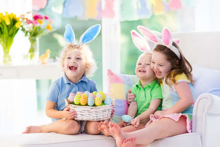 Kids celebrate Easter. Family, happy little girl, boy and baby in bunny ears on a couch. Children having fun on Easter egg hunt. Home decoration, pastel bunny banner, colorful Easter eggs and flowers