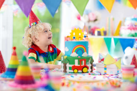 子供の誕生日パーティー カラフルなケーキにキャンドルを吹き消す子供 虹の旗の旗 風船で装飾された家 農場と輸送のテーマのお祝い 誕生日を祝う小さな男の子 パーティーフード の写真素材 画像素材 Image