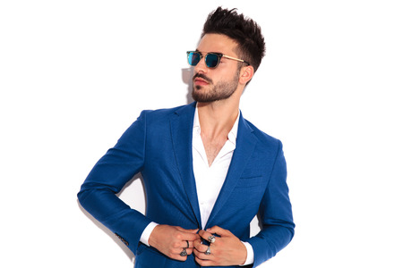 elegant man wearing sunglasses buttoning his suit and looks away to side on white background