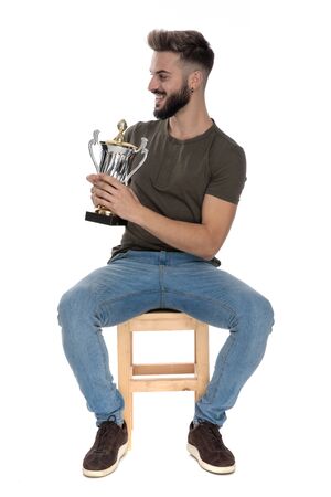 Cheerful casual man holding a trophy and laughing while sitting on a chair on white studio background