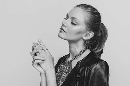 Side view of young woman in leather jacket looking to side, touching hands and posing on black and white background in studio