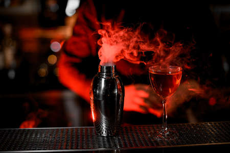 Smoking stainless steel shaker and tasty alcohol cocktail on a metal bar counter with bartender on background