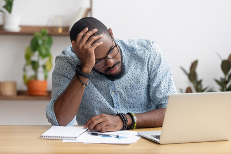 Tired depressed bored african businessman frustrated by business failure bankruptcy looking at laptop feel exhausted having headache, upset stressed black office worker worried about problem at work