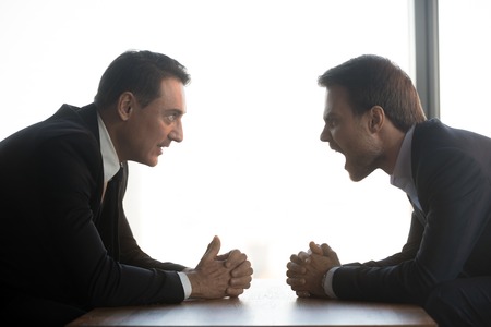 Mad businessmen sit at table opposite one another with clenched hands dispute scream at competitor, angry millennial men rivals in confrontation yell debate over desk, negotiate loud. rivalry concept Stock Photo